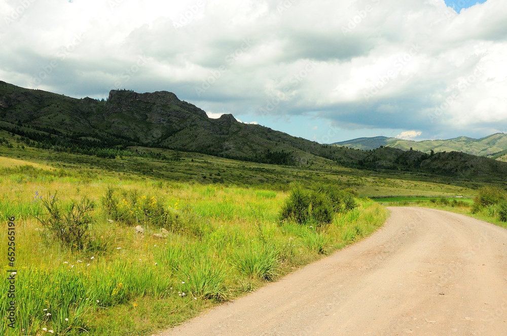 A gravel road turns through a valley at a high cliff under a cloudy summer sky.