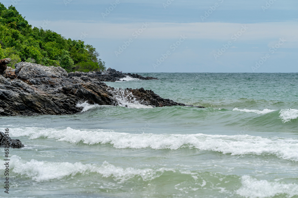 Low waves crash against the rocky shore of a tropical sea against a background of blue sky with white clouds.