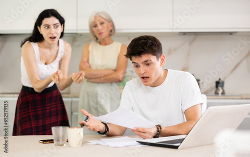 Young student guy does homework on laptop and swears with elderly mother and young girl sister. Conflict, quarrel, violation of personal boundaries