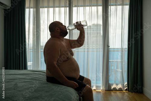 Extremely fat guy drinks lot of water, sits on bed, trying to cool off, alleviate serious condition, resist summer heat. Chubby, over obese, oversize man copes with dehydration due to profuse sweating