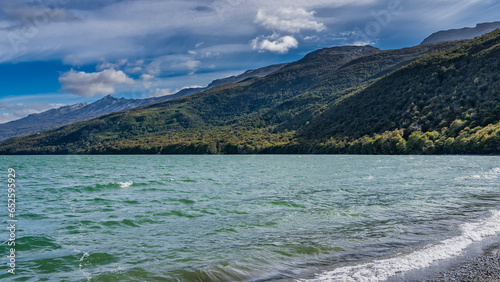 The waves of the emerald lake are foaming on the pebbly shore. Mountains covered with green forest, against a background of blue sky and clouds. Lago Roca. Argentina. Tierra del Fuego National Park.