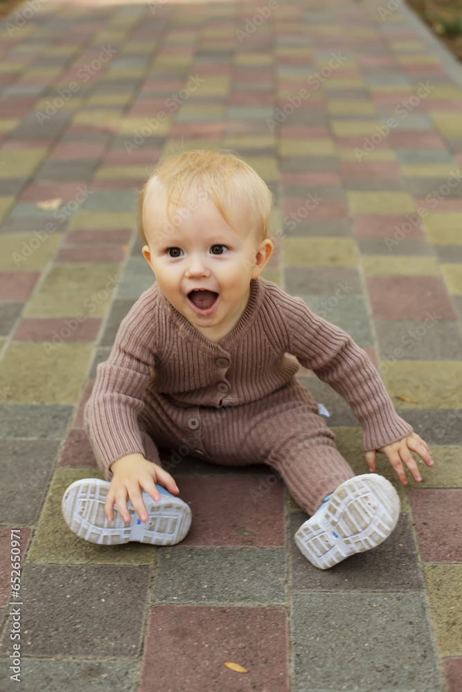Fototapeta premium a little boy in a brown jumpsuit sits happily on the sidewalk in the park