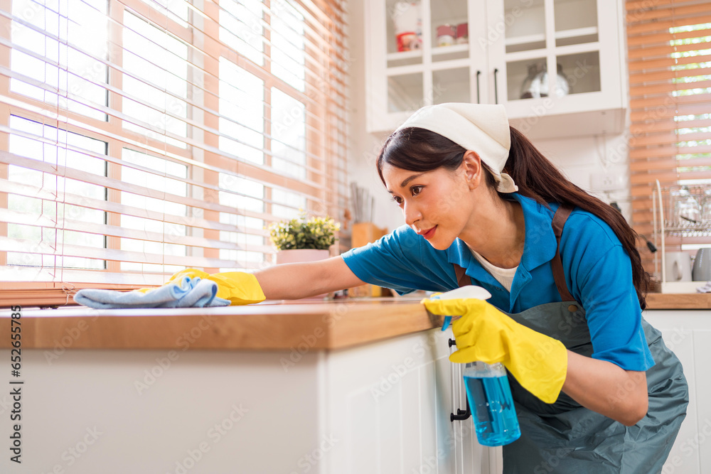 Professional Asian woman cleaning service wearing yellow rubber gloves, using a rag to wipe with spraying liquid detergent on the wooden top kitchen counter at home. Housekeeping cleanup, cleaner. Professional Asian woman cleaning service wearing yellow rubber gloves, using a rag to wipe with spraying liquid detergent on the wooden top kitchen counter at home. Housekeeping cleanup, cleaner.