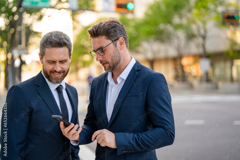 Two business men looking at messages on cellphone while walking in the street in downtown of city. Businessmen using smartphone mobile phone apps, texting message, browsing internet. Two business men looking at messages on cellphone while walking in the street in downtown of city. Businessmen using smartphone mobile phone apps, texting message, browsing internet.