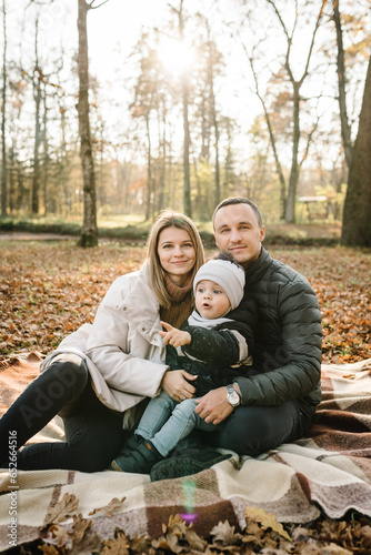 Mom, dad hugs son child sitting on blanket on yellow leaves in park. Autumn mood. Mother, father, kid have picnic in autumn forest in nature. Family spending time together at sunset. Autumn holiday.
