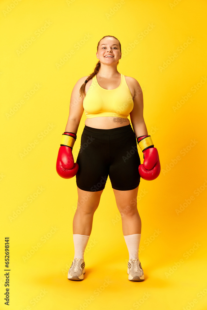 Full length portrait of attractive smiling plus size woman posing in sport outfit, with boxing gloves against yellow background. Full length portrait of attractive smiling plus size woman posing in sport outfit, with boxing gloves against yellow background.