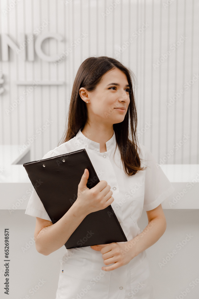 In front of the camera beautiful medical assistant posing to the camera at reception desk she smiling cute holding a map with documents in hands In front of the camera beautiful medical assistant posing to the camera at reception desk she smiling cute holding a map with documents in hands
