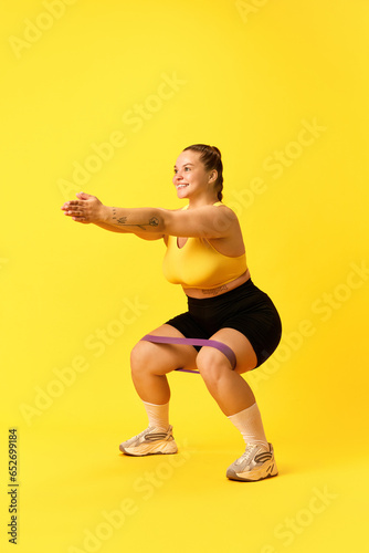 Fototapeta Naklejka Na Ścianę i Meble -  Full length portrait of young chubby sports woman in tracksuit squats with sport elastic bands for legs over yellow background.