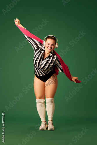 Overweight happy smiling woman dressed vintage sportwear doing exercises, with music in headphones with joy against green background.
