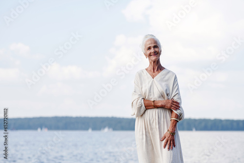 Portrait of elegant senior woman smiling at camera while standing outdoors against the lake