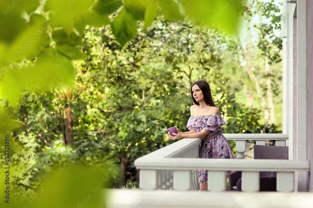 A girl in a lilac dress stands on the balcony of her house. She is thoughtful, holding a mug of tea in her hands. Season of the year is summer.