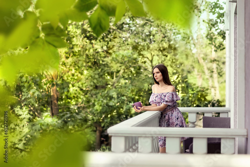 A girl in a lilac dress stands on the balcony of her house. She is thoughtful, holding a mug of tea in her hands. Season of the year is summer.