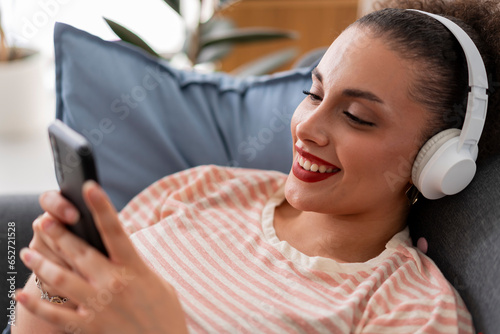 Cheerful young female in casual clothes smiling and listening to music in wireless headphones and browsing mobile phone while lying on sofa at home photo
