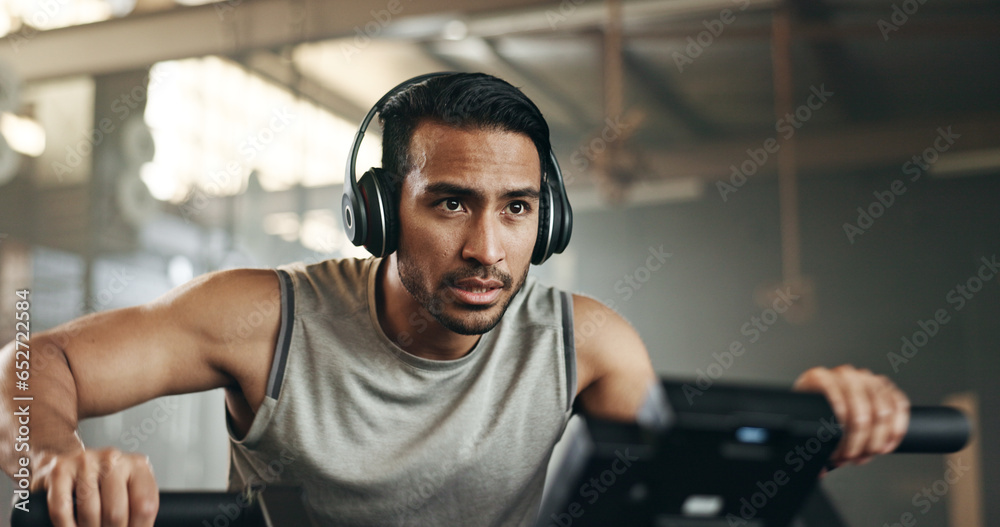 Asian man, headphones and cycling at gym on machine and listening to music in sports workout or exercise. Serious male person or athlete training on bicycle machine or equipment for healthy cardio Asian man, headphones and cycling at gym on machine and listening to music in sports workout or exercise. Serious male person or athlete training on bicycle machine or equipment for healthy cardio