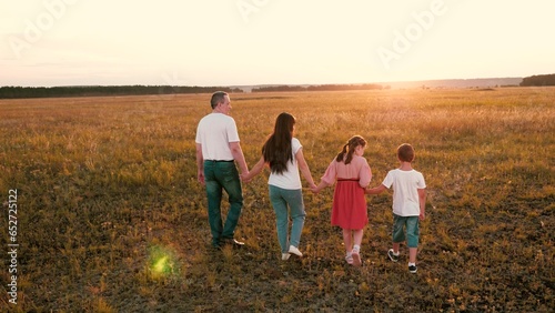 Loving parents hold hands of cute children walking across evening field