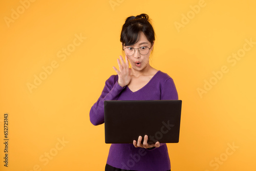 A confident and excite female entrepreneur wearing a purple shirt and eyeglasses, with a laptop, portraying a strong presence in the business world in a studio portrait against a yellow background.