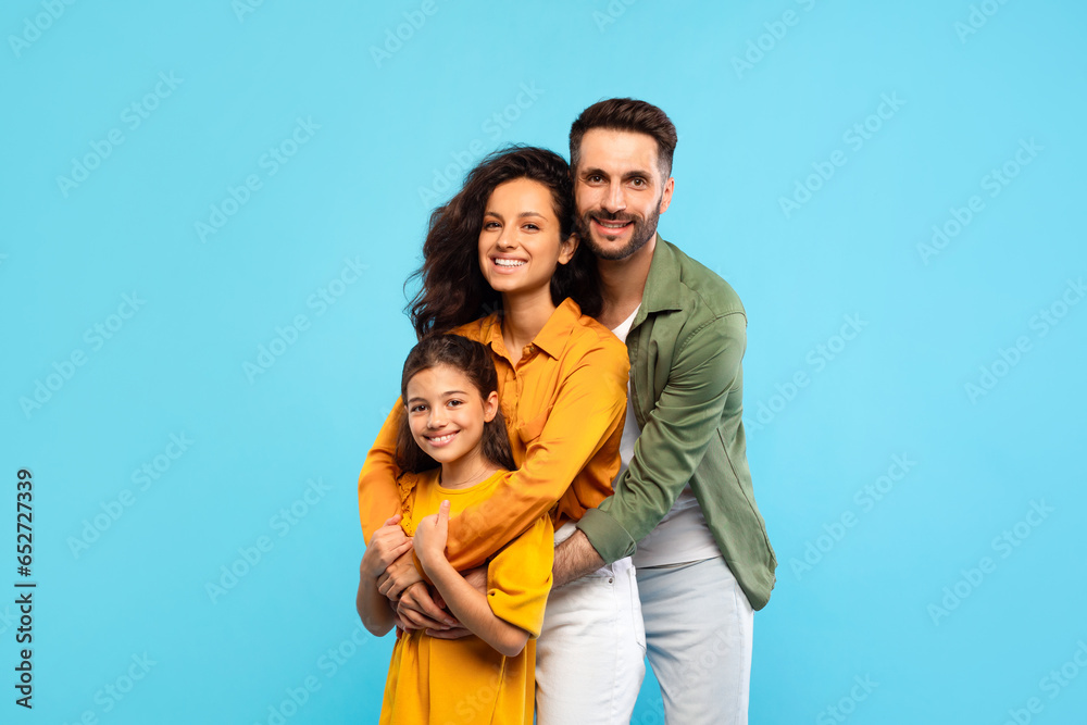 Happy caucasian family of three with daughter embracing and smiling at camera, posing on blue background Happy caucasian family of three with daughter embracing and smiling at camera, posing on blue background
