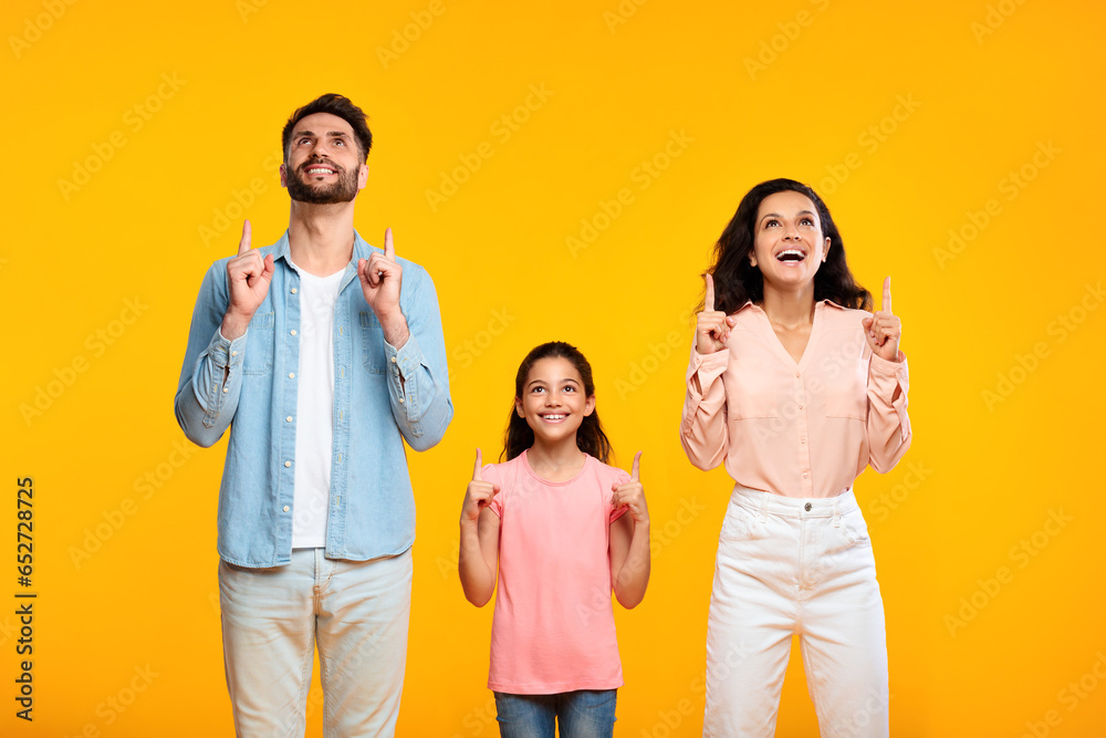 Joyful european family of three looking and pointing fingers up, posing standing over yellow background, free space Joyful european family of three looking and pointing fingers up, posing standing over yellow background, free space