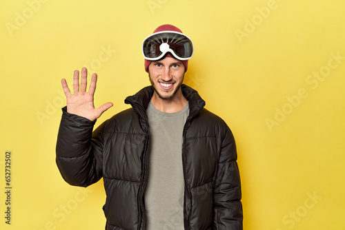 Man in winter coat with snow goggles on yellow smiling cheerful showing number five with fingers.