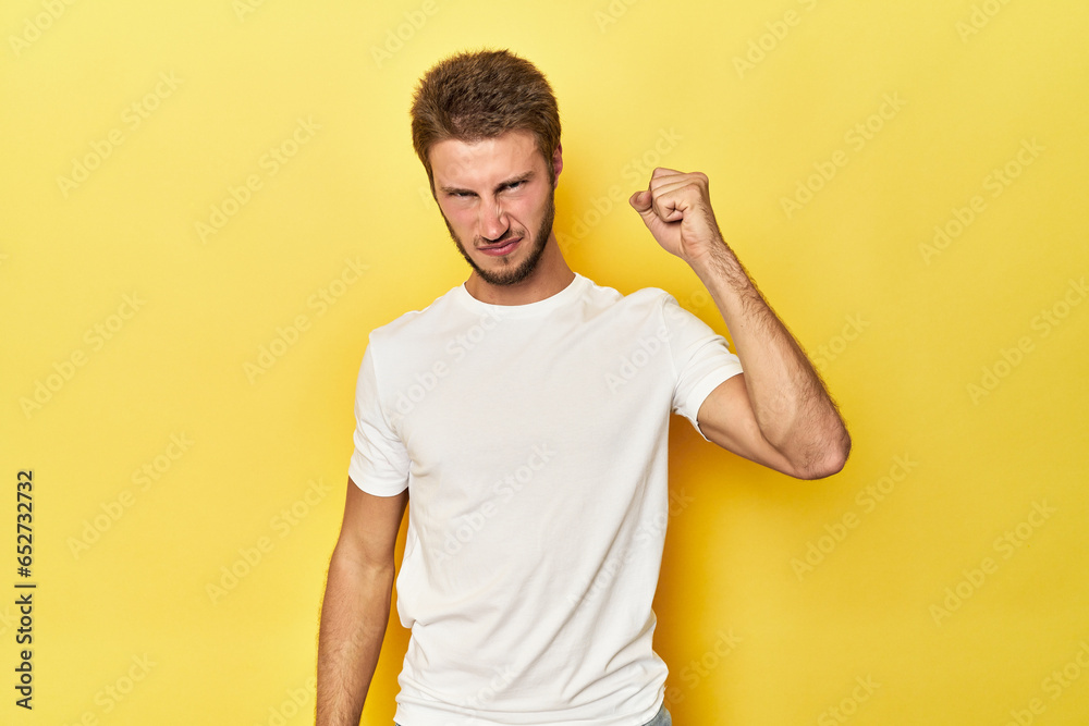 Young Caucasian man on a yellow studio background showing fist to camera, aggressive facial expression. Young Caucasian man on a yellow studio background showing fist to camera, aggressive facial expression.