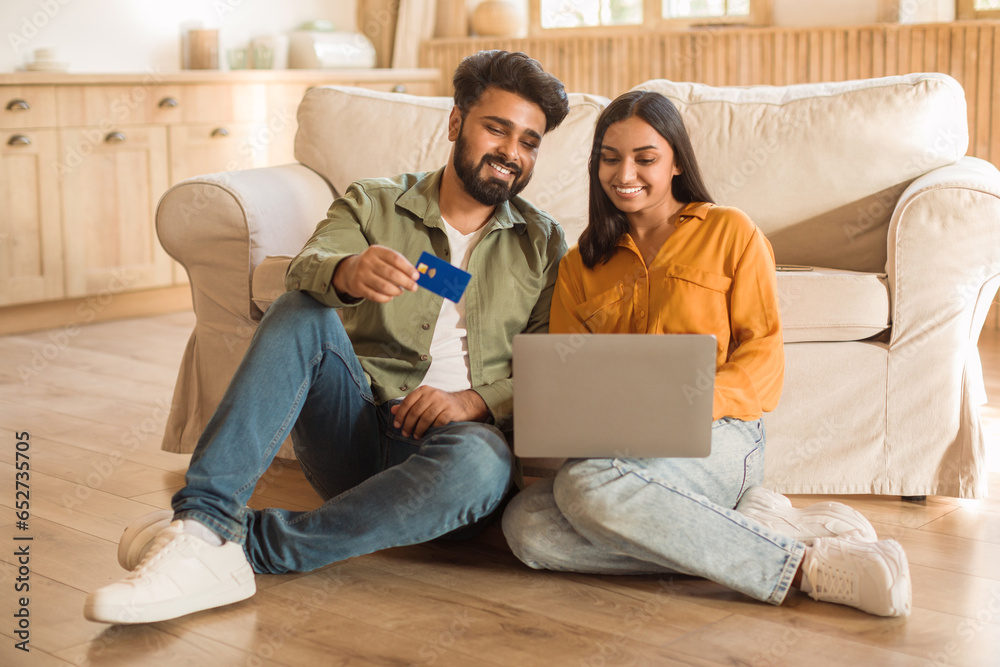 Happy indian spouses shopping online via laptop and using credit card, sitting on floor at home interior, copy space Happy indian spouses shopping online via laptop and using credit card, sitting on floor at home interior, copy space