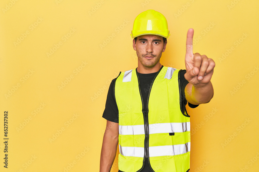 Latino worker in yellow vest and helmet showing number one with finger. Latino worker in yellow vest and helmet showing number one with finger.
