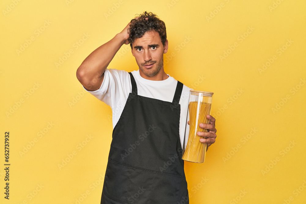 Young latino man cook with pasta jar on yellow being shocked, she has remembered important meeting. - obrazy, fototapety, plakaty 