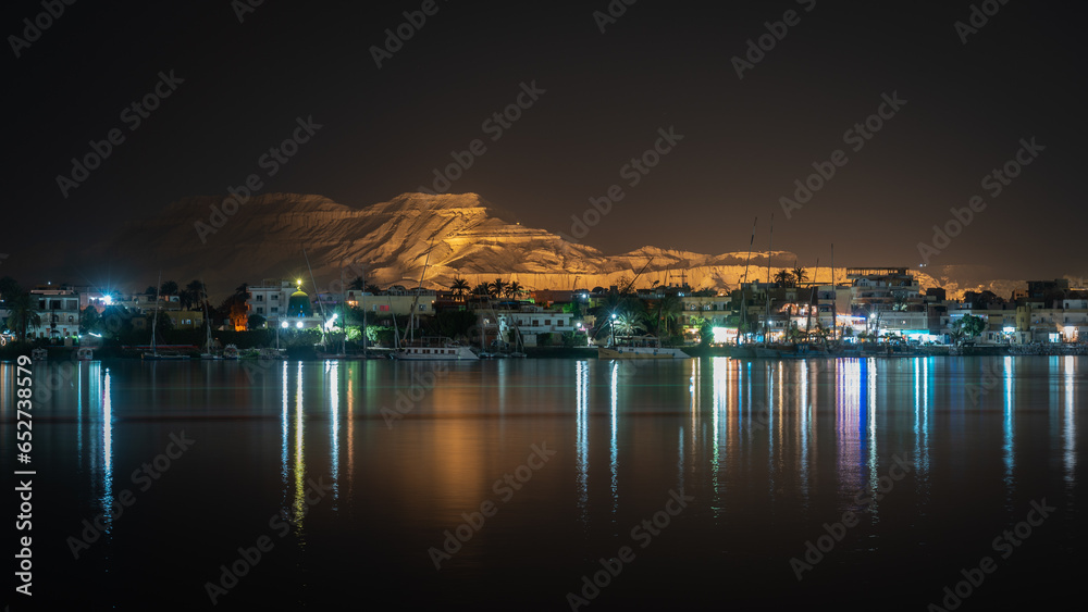 Night image of the illuminated Valley of the Kings and its reflection in the water of the Nile River as it passes through Luxor, Egypt.
