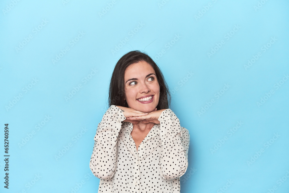 Young caucasian woman on blue backdrop keeps hands under chin, is looking happily aside. Young caucasian woman on blue backdrop keeps hands under chin, is looking happily aside.