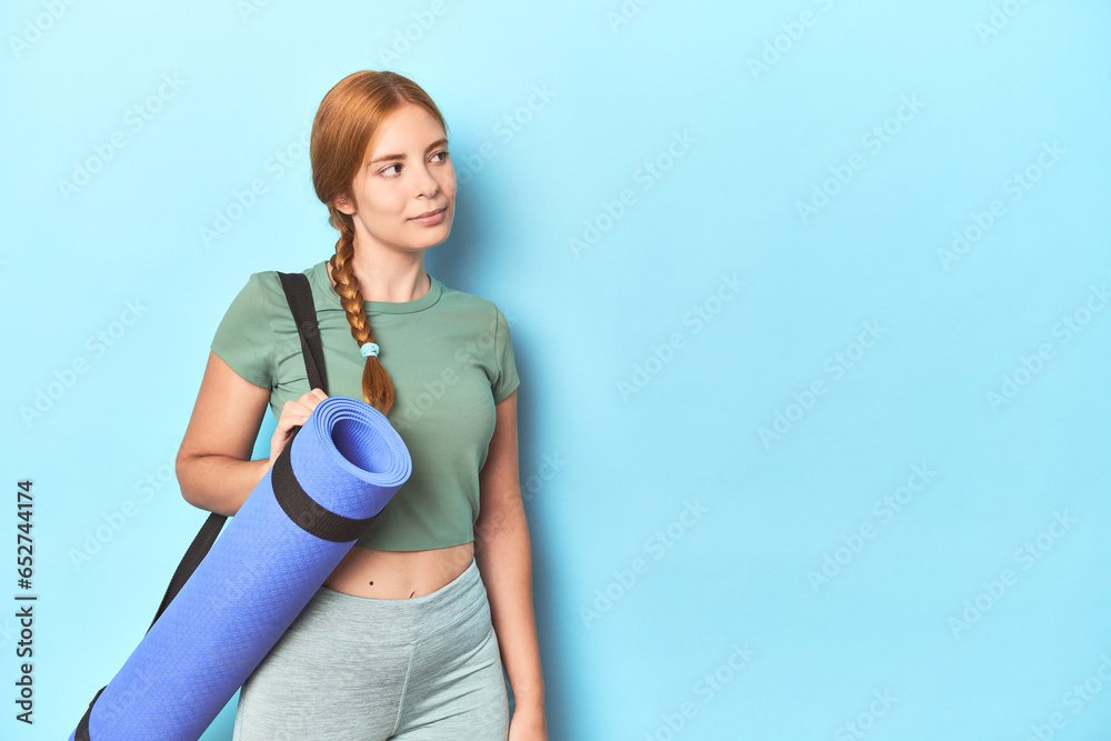Sporty redhead woman carrying yoga mat in blue studio background Sporty redhead woman carrying yoga mat in blue studio background
