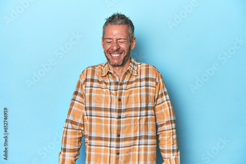 Middle-aged caucasian man on blue backdrop laughs and closes eyes, feels relaxed and happy.