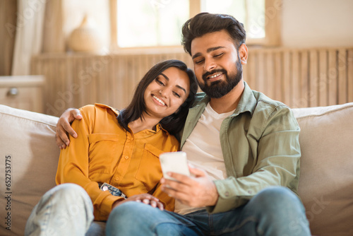 Happy loving hindu couple using cellphone, having video call or browsing internet, man embracing wife, sitting on sofa