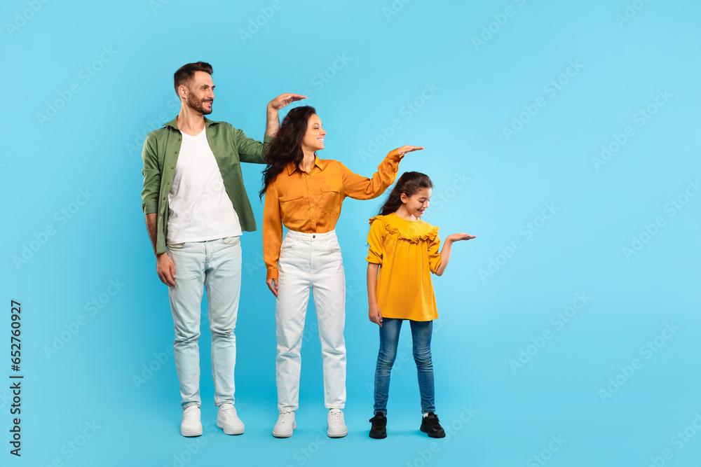 Family hierarchy. Young parents and daughter measuring each other's height with hand, posing over blue background - obrazy, fototapety, plakaty 