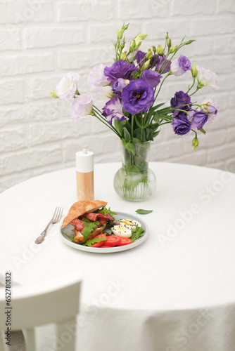 Breakfast of croissant burger with fries ham, boiled eggs on round white table with vase of bouquet of eustomas on light bricks background.