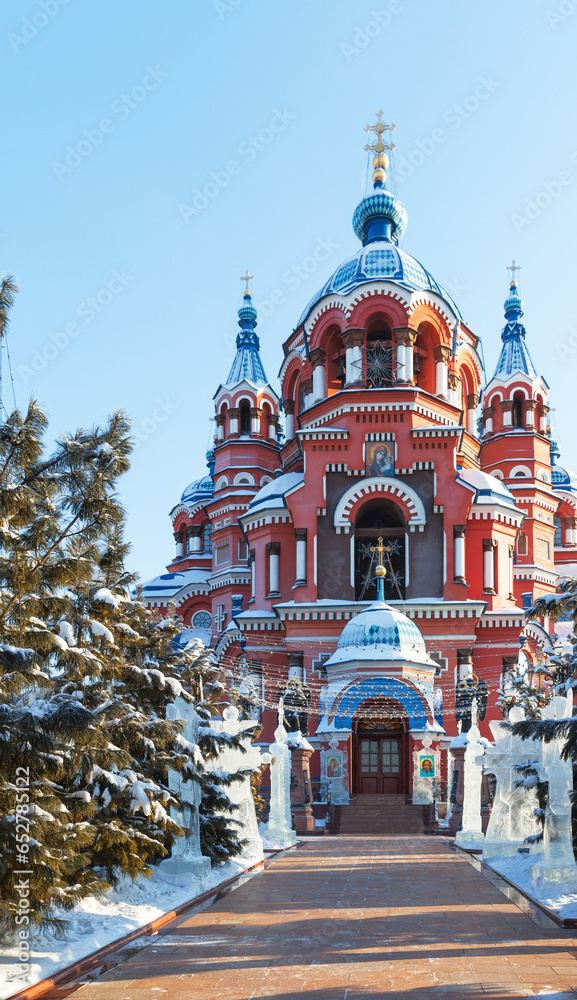 Irkutsk in winter holidays. City landmark - Cathedral of Kazan Icon of Mother of God during Christmas. Сourtyard of cathedral is decorated with ice sculptures and Christmas tree - obrazy, fototapety, plakaty 