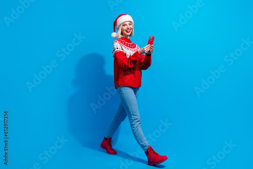 Full length photo of shiny adorable woman santa elf wear ornament pullover walking texting modern gadget isolated blue color background