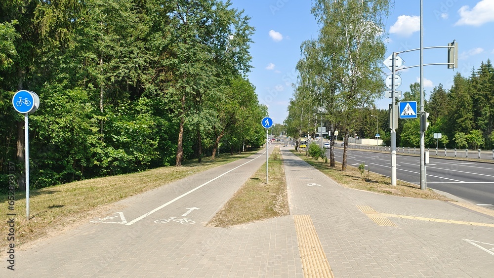 Tile-paved bike and pedestrian paths, marked with signs and pavement markings, are located between the park's trees and the roadway of the city street where cars travel. Summer sunny weather