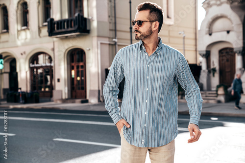 Portrait of handsome confident stylish hipster lambersexual model. Sexy modern man dressed in blue shirt and trousers. Fashion male posing on street background in Europe city at sunset. In sunglasses