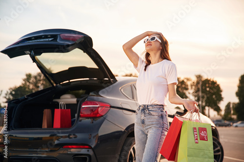 Against the car with trunk opened. Beautiful woman in casual clothes is holding shopping bags, outdoors