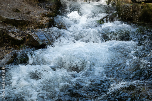 A stormy stream of a mountain river flows over large and small stones, white waves, strong current, small waterfalls, swirls, dark water