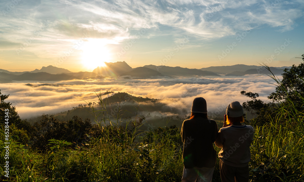 Two female tourist friends sit and admire the sea of mist, walk to the top of the mountain and watch the mountain view of the sunrise in the morning.