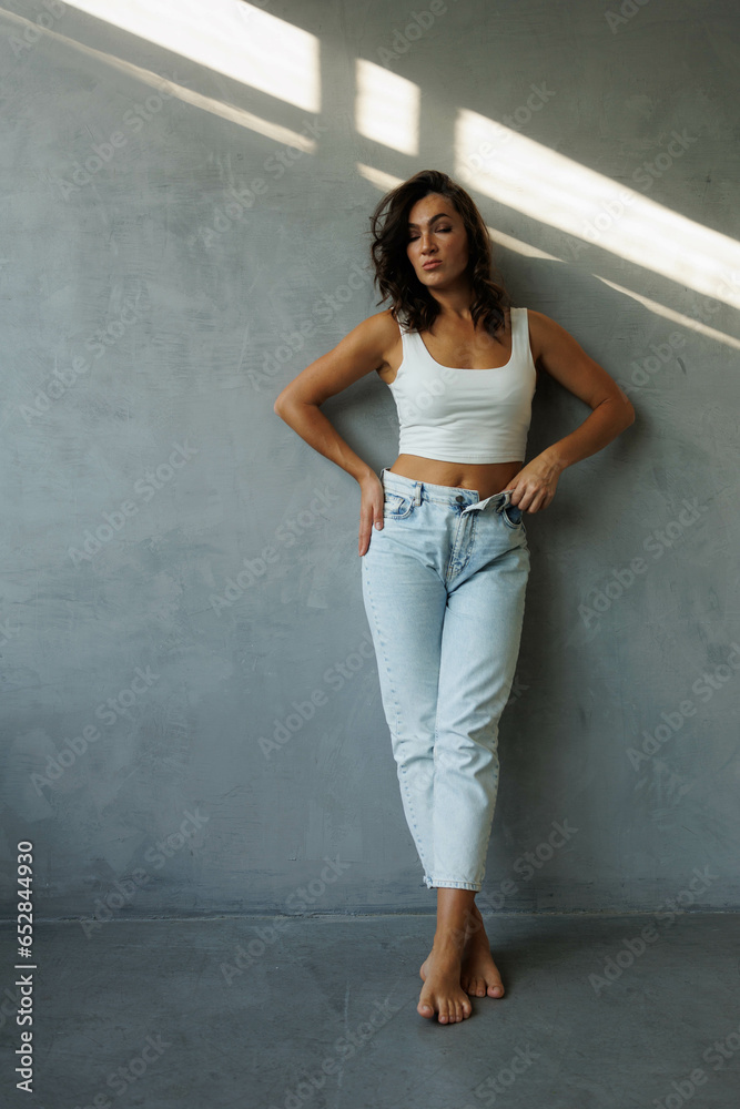Beautiful brunette woman in white top and blue jeans, standing. Girl smile, happy. Portrait of young pretty woman. Complicated sunlight with shadows. White background. Light from window, portrait. Beautiful brunette woman in white top and blue jeans, standing. Girl smile, happy. Portrait of young pretty woman. Complicated sunlight with shadows. White background. Light from window, portrait.