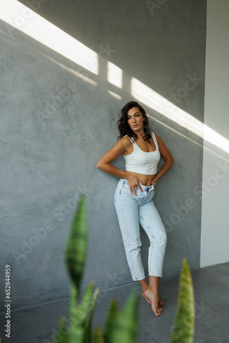 Beautiful brunette woman in white top and blue jeans, standing. Girl smile, happy. Portrait of young pretty woman. Complicated sunlight with shadows. White background. Light from window, portrait.