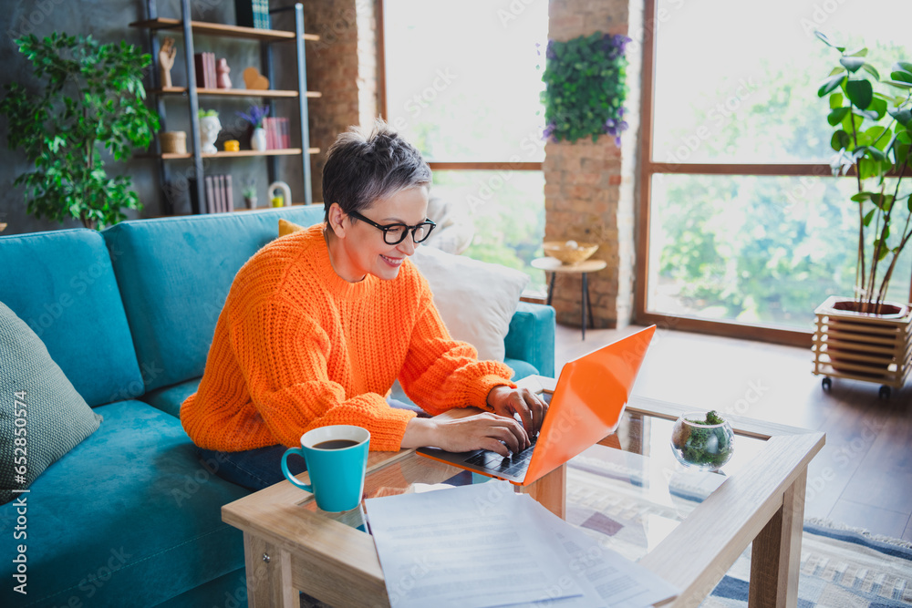 Photo of good mood cheerful mature lady dressed orange sweater typing apple samsung modern device indoors house room Photo of good mood cheerful mature lady dressed orange sweater typing apple samsung modern device indoors house room