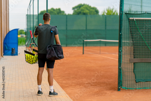 young professional player coach standing on outdoor tennis court before starting game training with racket and basket of tennis balls photo