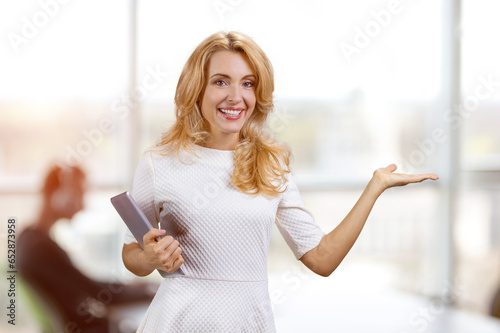 Smiling blonde mature woman with tablet advertising copy space. Office interior in the background.