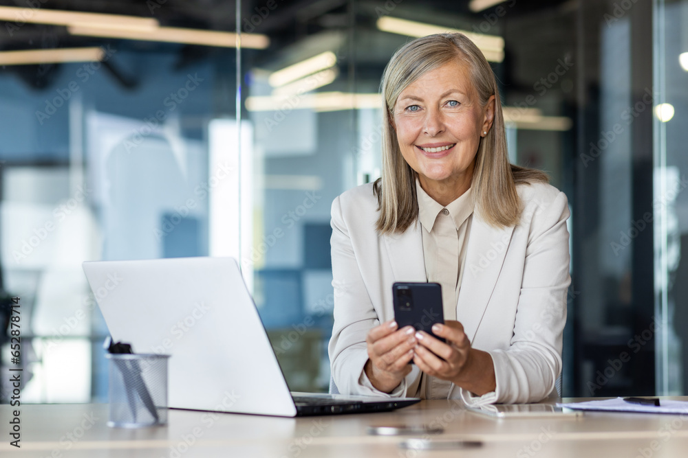 Portrait of mature gray-haired businesswoman inside office at workplace, woman boss smiling and looking at camera, holding phone in hands using smartphone app, sitting at table with laptop. Portrait of mature gray-haired businesswoman inside office at workplace, woman boss smiling and looking at camera, holding phone in hands using smartphone app, sitting at table with laptop.