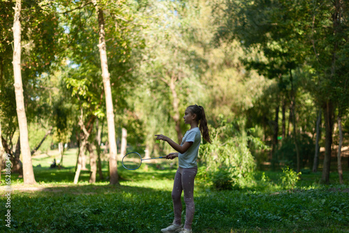 little cute caucasian girl, playing badminton in a summer park, surrounded by green trees