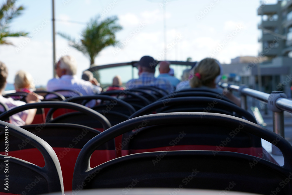 Obraz premium Double decker hop on hot off bus with open roof with some defocused passengers in rear view. On the background there is a palm tree against clear sky. Tourist bus cutout with focus on the foreground.
