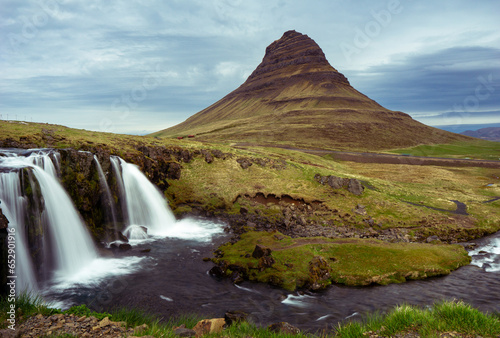 Clouds embrace Kirkjufell, where waterfalls dance and the mountain stands, shrouded in nature's mystique.